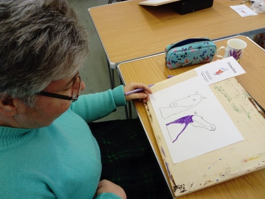 Woman in blue jumper is sat in her wheelchair at a wooden desk. She is taking part in an Ecas art activity and is painting The Kelpies.