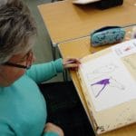 Woman in blue jumper is sat in her wheelchair at a wooden desk. She is taking part in an Ecas art activity and is painting The Kelpies.